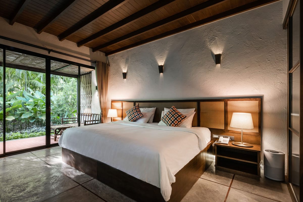 A spacious bedroom with a large bed, wooden headboard, bedside lamps, and a view of lush greenery through large windows.