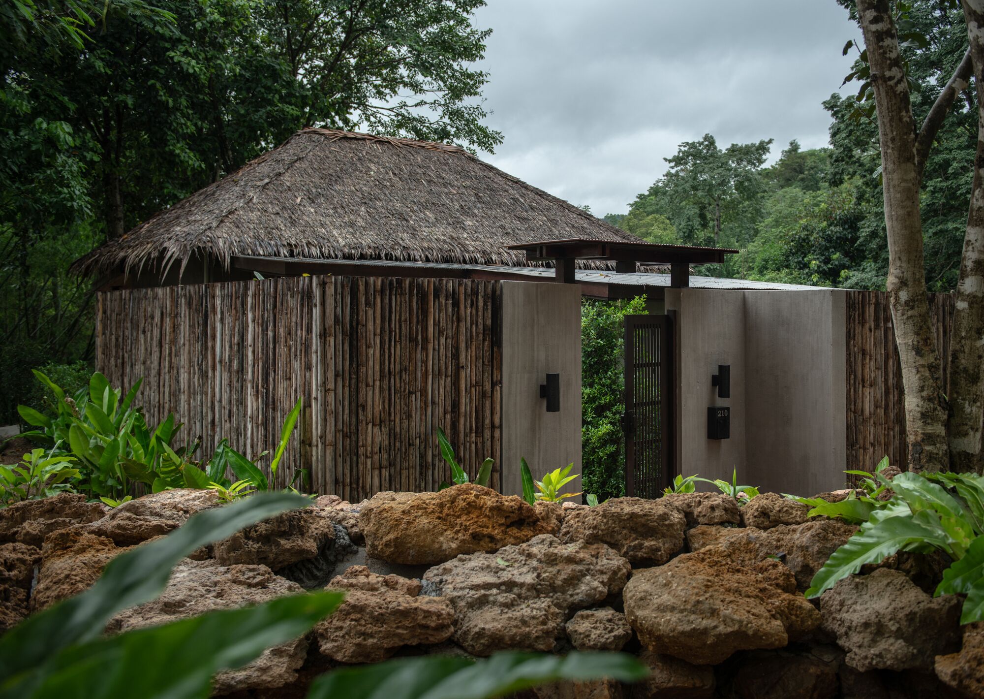 A rustic hut with a thatched roof, surrounded by lush greenery and a rocky foreground.
