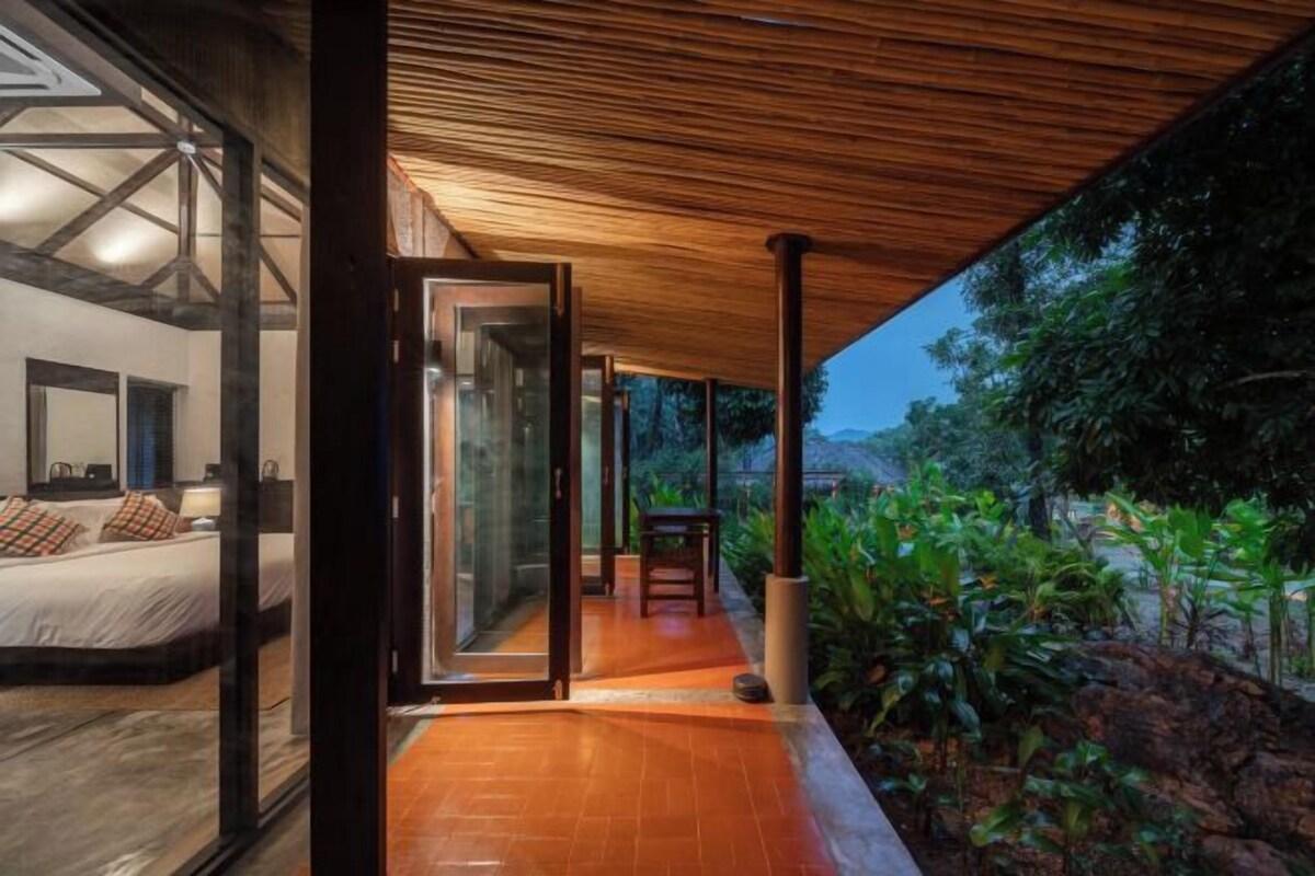A modern hotel room with a large bed, a glass door leading to a balcony, and a view of the surrounding greenery.