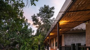 A covered outdoor area with a wooden ceiling, surrounded by lush greenery and a pathway leading to a building entrance. - Phapok Eco Resort (Suan Phueng)