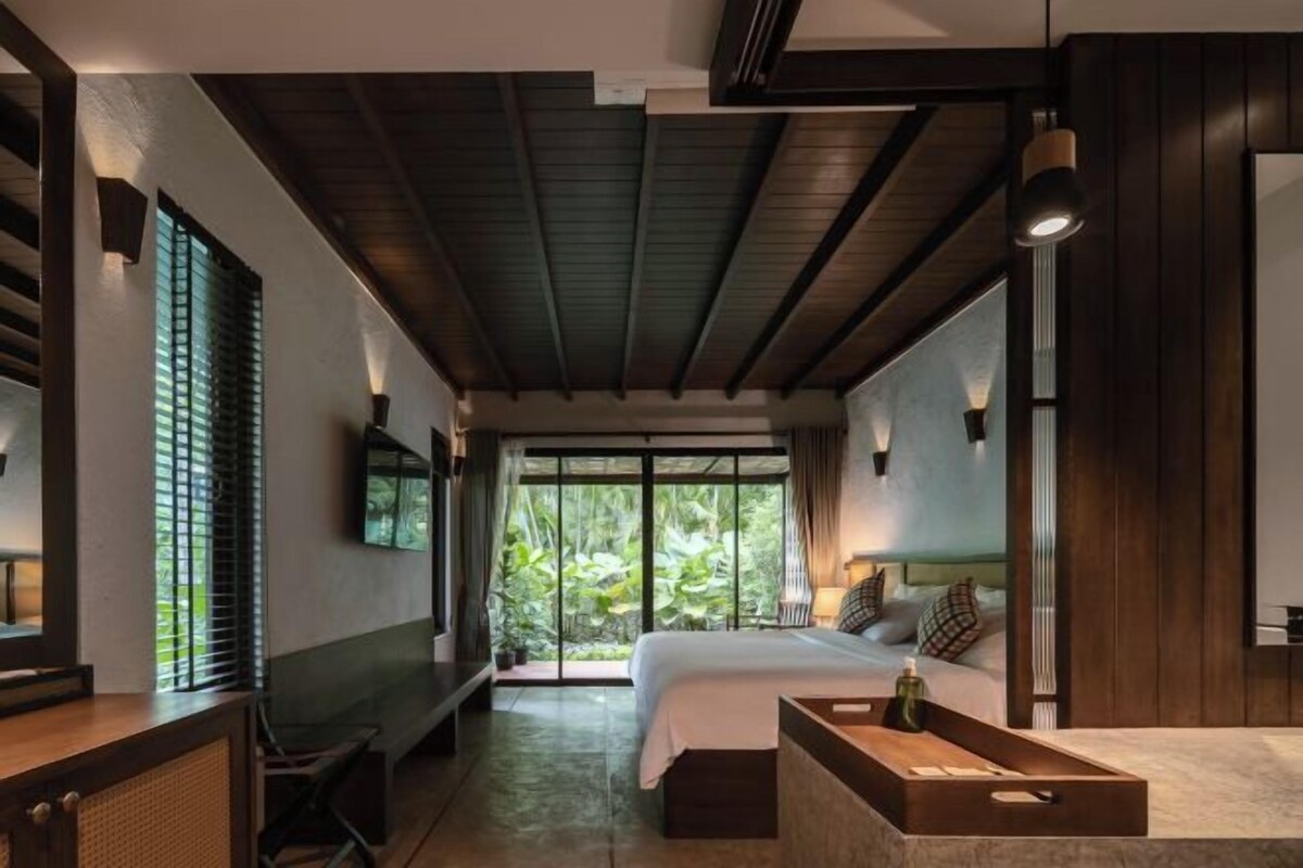 A modern bedroom with a large bed, a desk, and a view of greenery through the window.