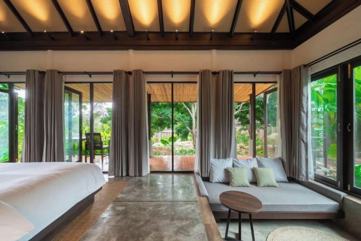 A modern bedroom with a large bed, a sofa, and a small table. There are curtains, a view of greenery outside, and a ceiling with exposed beams.
