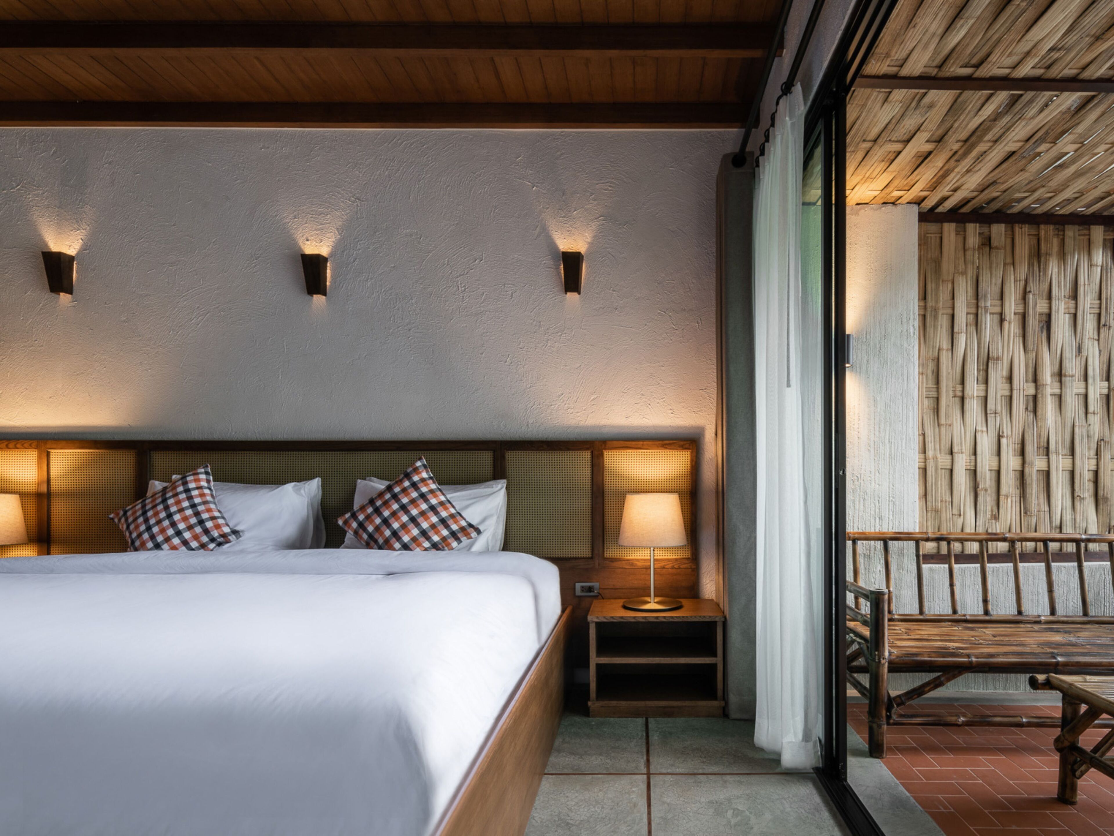 Une chambre à coucher avec un grand lit, deux tables de chevet avec des lampes, une tête de lit en bois et une vue sur un mur de bambou à l’extérieur.