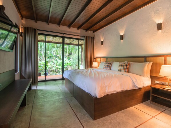 A bedroom with a large bed, a bedside table, a TV, and a view of greenery through the window.