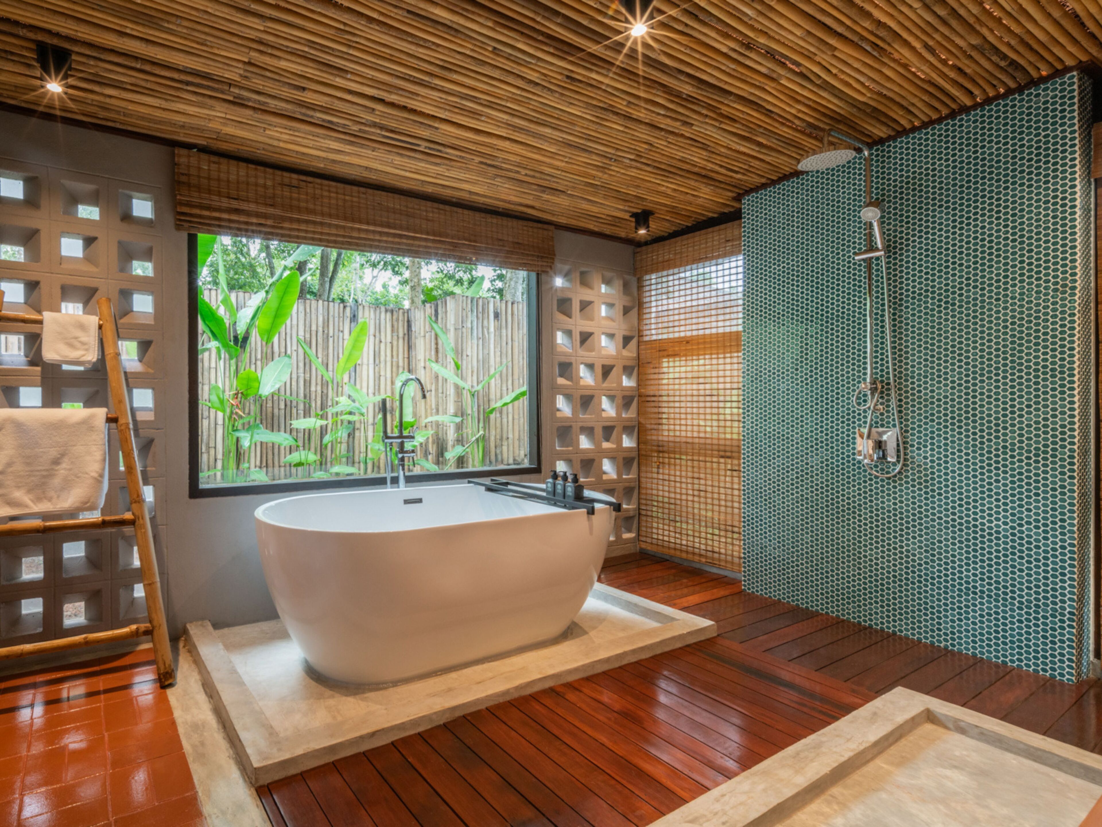 A modern bathroom with a freestanding bathtub, a window with a view of greenery, and a shower area with a patterned wall.