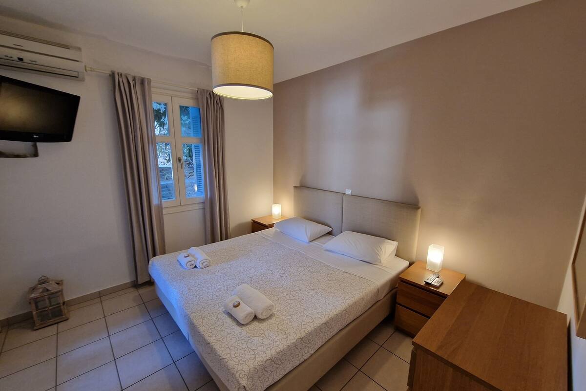 A neatly made bed with a patterned bedspread and two pillows, a bedside table with a lamp, a wall-mounted TV, and a window with curtains.