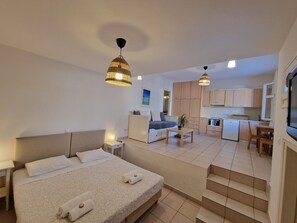 Quarto de hotel moderno com cama grande, mesas de cabeceira, escrivaninha pequena, sofá, mesa de jantar e área de cozinha.
