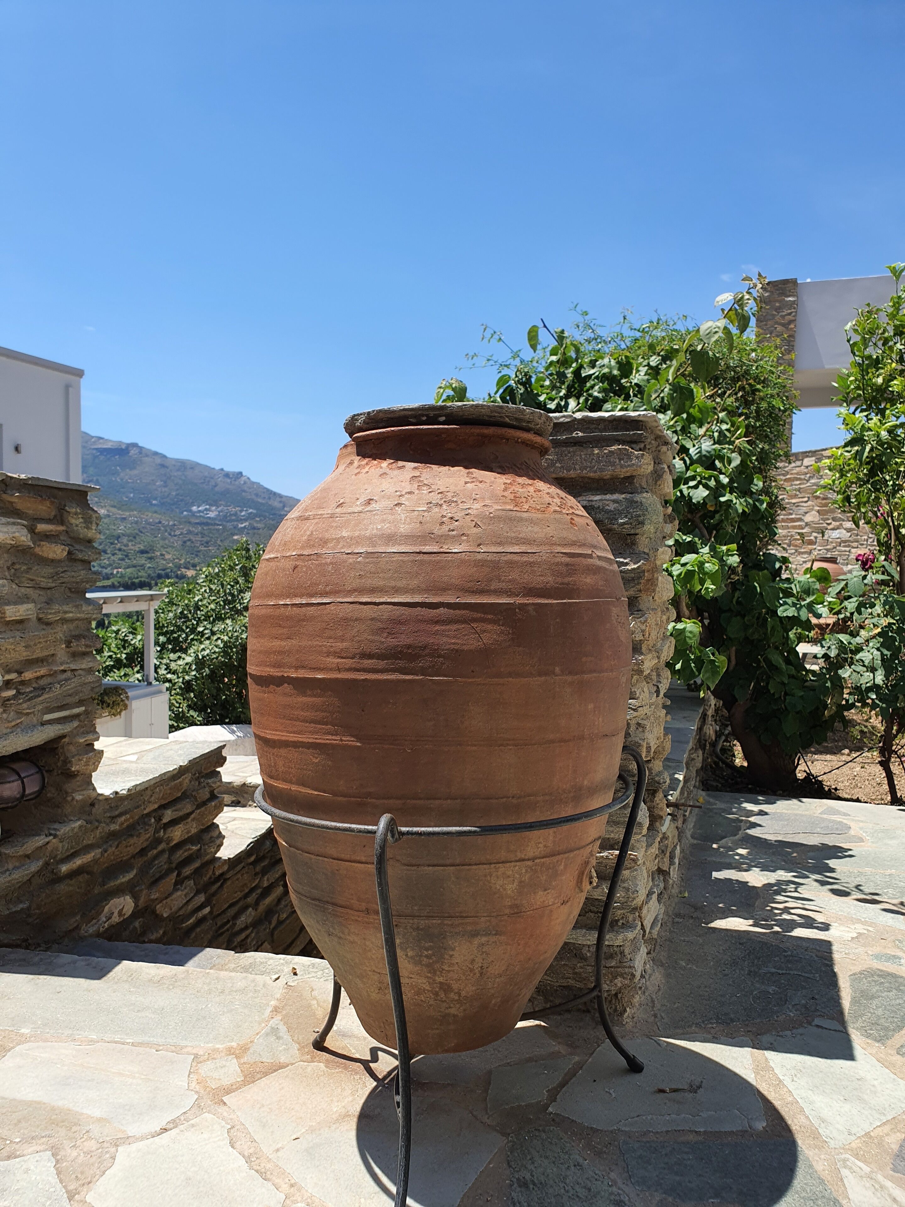 Una gran maceta de terracota en un patio de piedra, con vistas a montañas y un edificio blanco al fondo.