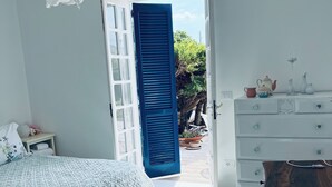 Ein Schlafzimmer mit Bett, Schminktisch und Schreibtisch. Ein Fenster mit blauen Fensterläden und Blick nach draußen.
