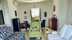Ein Wohnzimmer mit einem gemusterten Sofa, einem grünen Sessel, einem weißen Sofa, einem Esstisch mit Stühlen und einer kleinen Kommode.