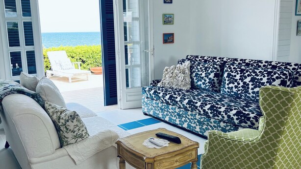 Ein Wohnzimmer mit einem weißen Sofa, einem gemusterten Sofa, einem grünen Sessel und einem kleinen Holztisch. Es gibt ein großes Fenster mit Blick aufs Meer.