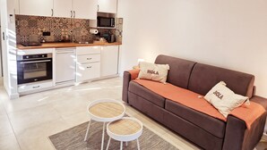 A modern kitchen with white cabinets, a microwave, and an oven. There is a brown sofa with two pillows, one with "HOLA" on it, and two round stools with woven seats. - A Casuccia di Mammò (Oletta)