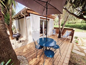 Une terrasse en bois avec une table et des chaises bleues, un barbecue et un espace salon couvert avec une table et des chaises.