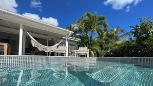 Une piscine d’un bleu éclatant, bordée de carreaux, entourée de palmiers et agrémentée d’un espace de repos couvert avec des chaises.