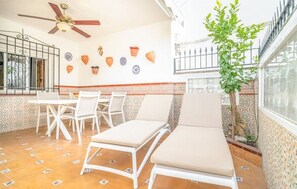 Pátio ensolarado com ventoinha no teto, cadeiras de descanso brancas, uma mesa e azulejos decorativos.