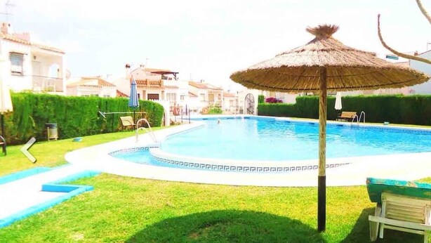 A swimming pool with a thatched umbrella, surrounded by a well-maintained lawn and a few lounge chairs. - Holiday Home "Casa Colibri 2 Beach" with Shared Pool & Wi-Fi (Nerja)