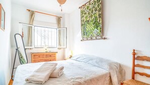 A bedroom with a bed, a wooden dresser, a chair, a window with curtains, and a tapestry on the wall. - Holiday Home "Casa Colibri 2 Beach" with Shared Pool & Wi-Fi (Nerja)