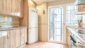 A kitchen with wooden cabinets, a white refrigerator, a sink, and a window with metal bars. - Holiday Home "Casa Colibri 2 Beach" with Shared Pool & Wi-Fi (Nerja)