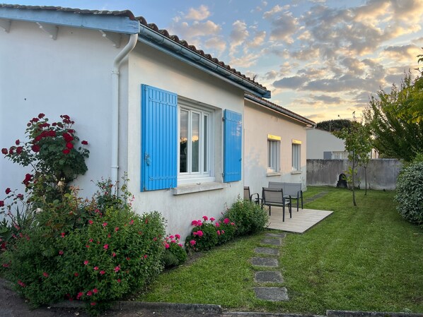 Une maison à l’extérieur blanc, avec des volets bleus, un petit jardin agrémenté de fleurs roses et un chemin de pierres menant à un espace aménagé pour s’asseoir.