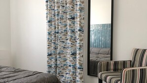 A bedroom with a bed, a striped armchair, a teal side table, a mirror, and a curtain with a fish pattern. - Holiday Home 'Picotelle' with Private Terrace, Private Garden and Wi-Fi (Le Château d'Oléron)