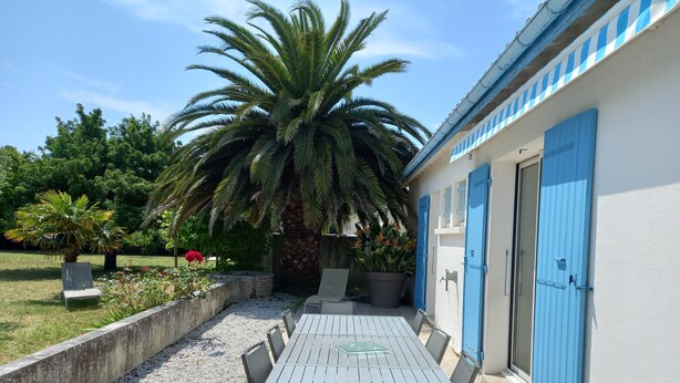 A patio with a table and chairs, a palm tree, and a building with blue doors. - Holiday Home 'Picotelle' with Private Terrace, Private Garden and Wi-Fi (Le Château d'Oléron)
