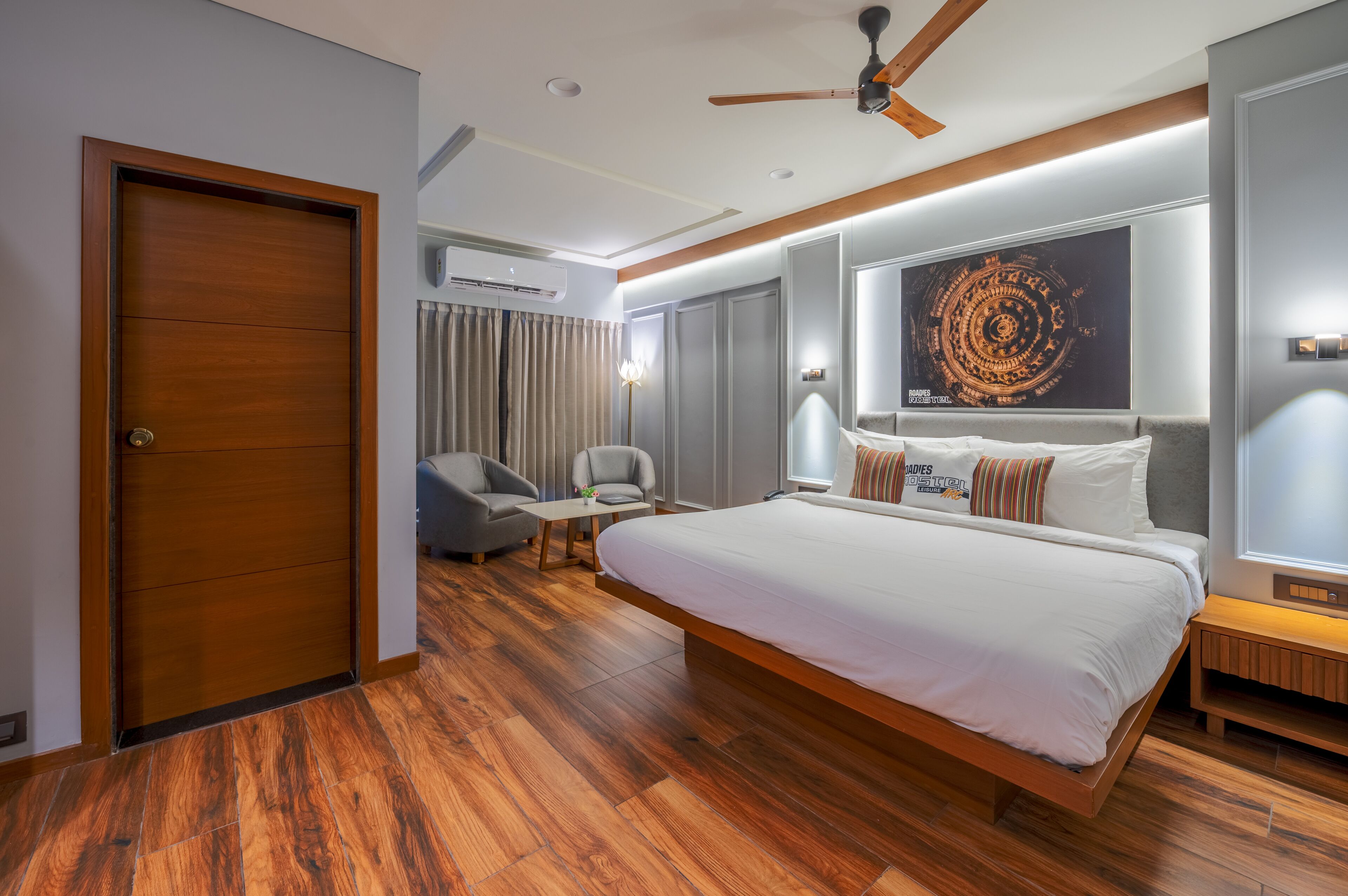 Una habitación de hotel moderna con una cama grande, un ventilador de techo, un área de descanso con una mesa y sillas, y una obra de arte de gran tamaño en la pared.
