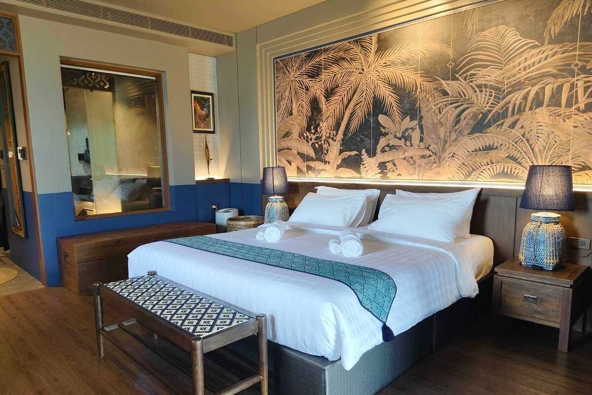 Een hotelkamer met een groot bed, een nachtkastje, een bankje, een spiegel en een wanddecoratie met een tropisch thema.