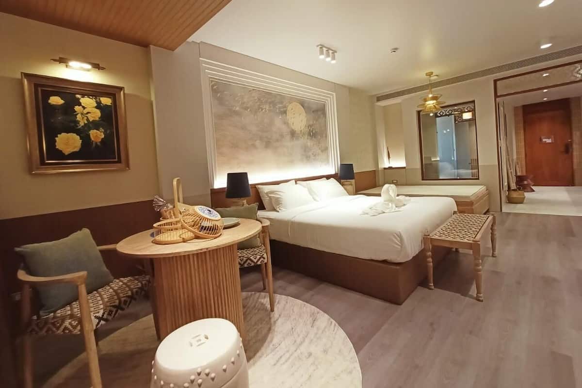 Een hotelkamer met een groot bed, een ronde tafel, een stoel en een klein krukje. Er is een groot raam met jaloezieën, een spiegel en een deur naar een andere kamer.