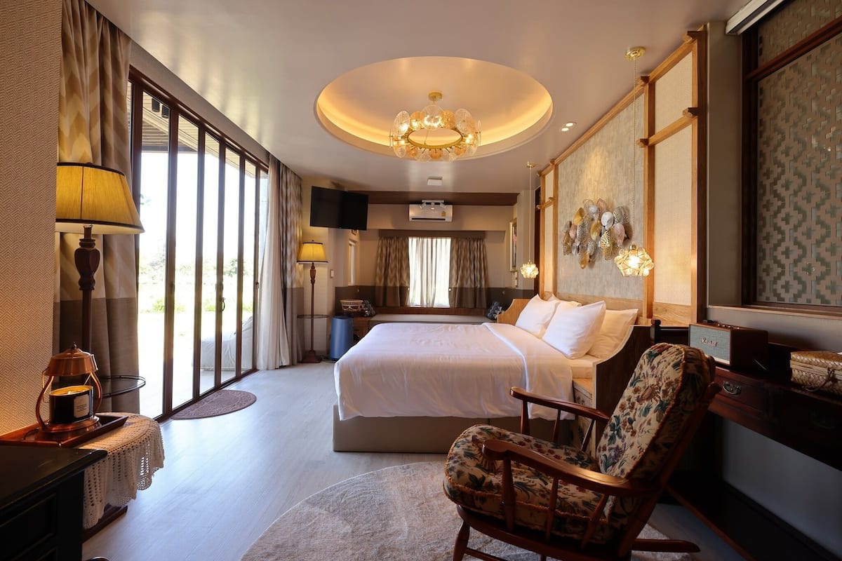 Ruime hotelkamer met een groot bed, een zithoek met een fauteuil en uitzicht door de grote ramen.