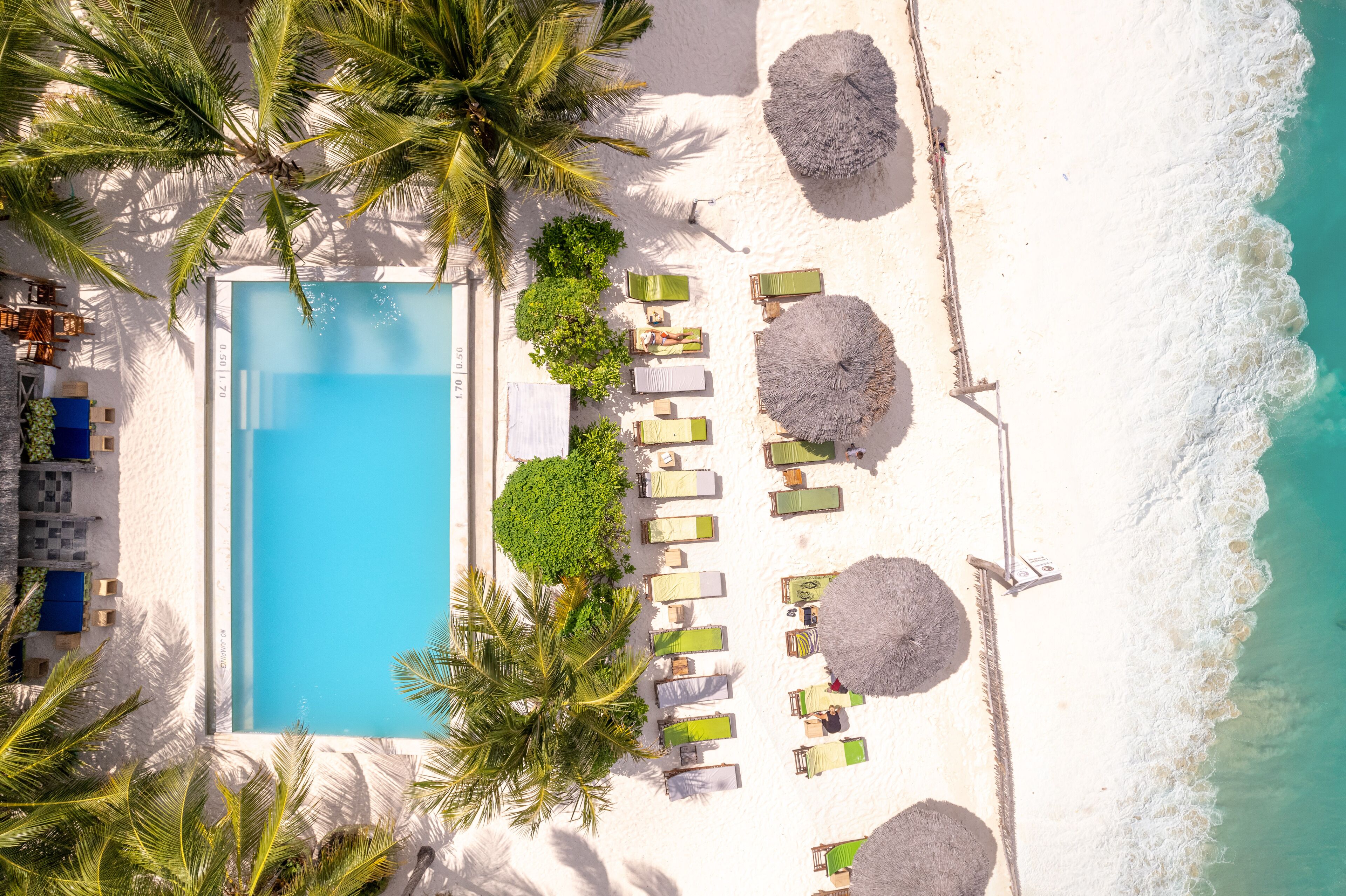 An aerial view of a beachside resort with a swimming pool, sun loungers, and palm trees.