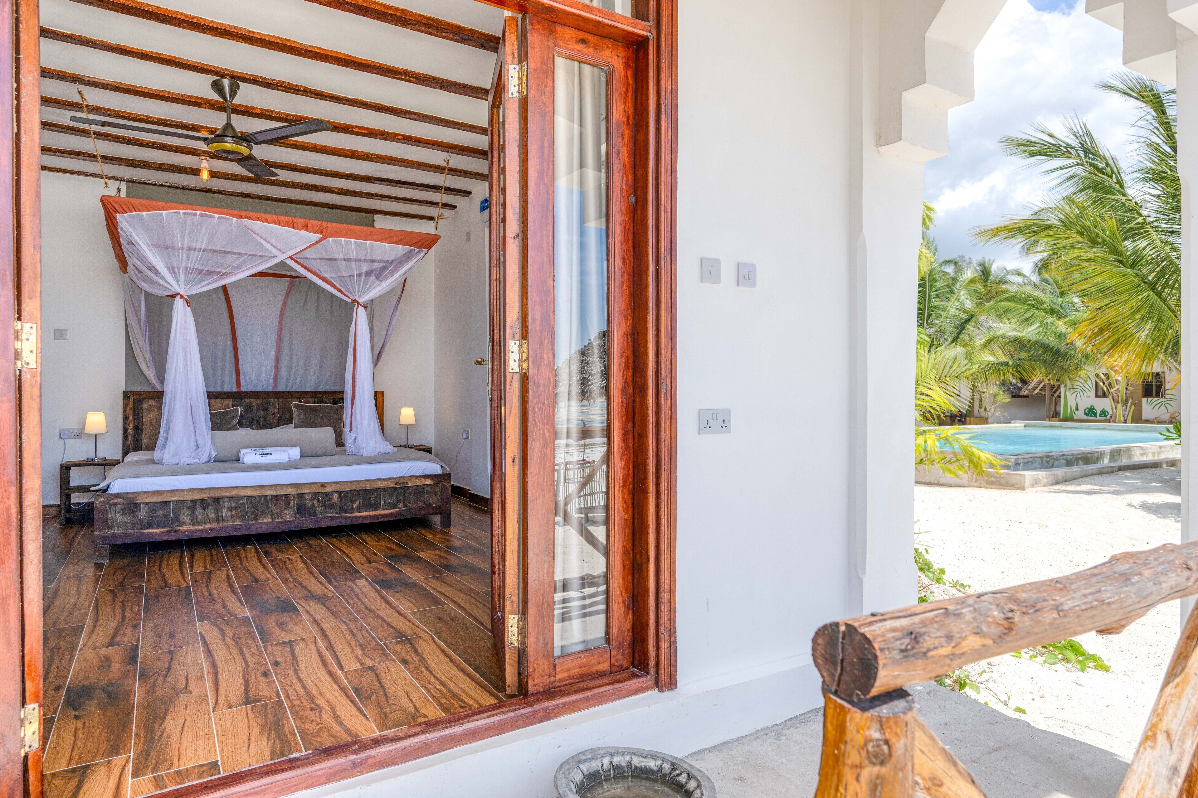 Een slaapkamer met een houten vloer, een bed met baldakijn en uitzicht op een zwembad en palmbomen.