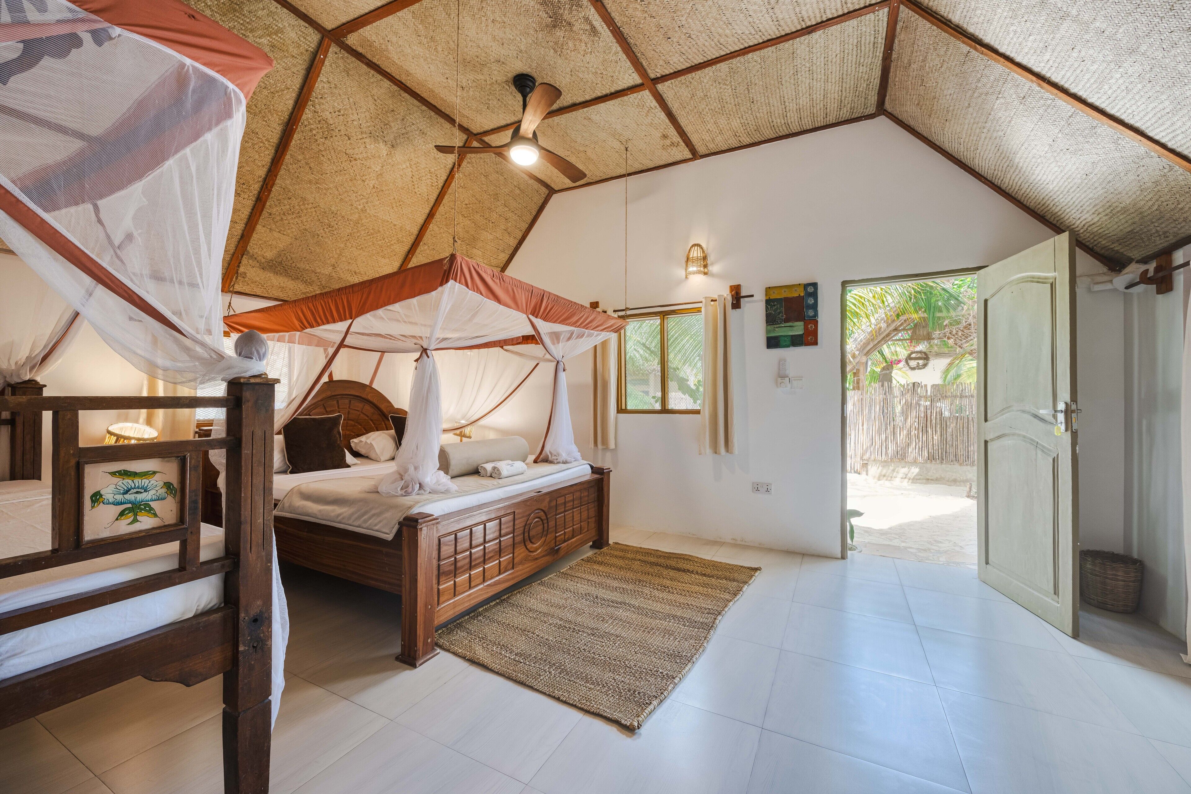 A bedroom with a wooden bed, a mosquito net, a ceiling fan, and a view of the outside.