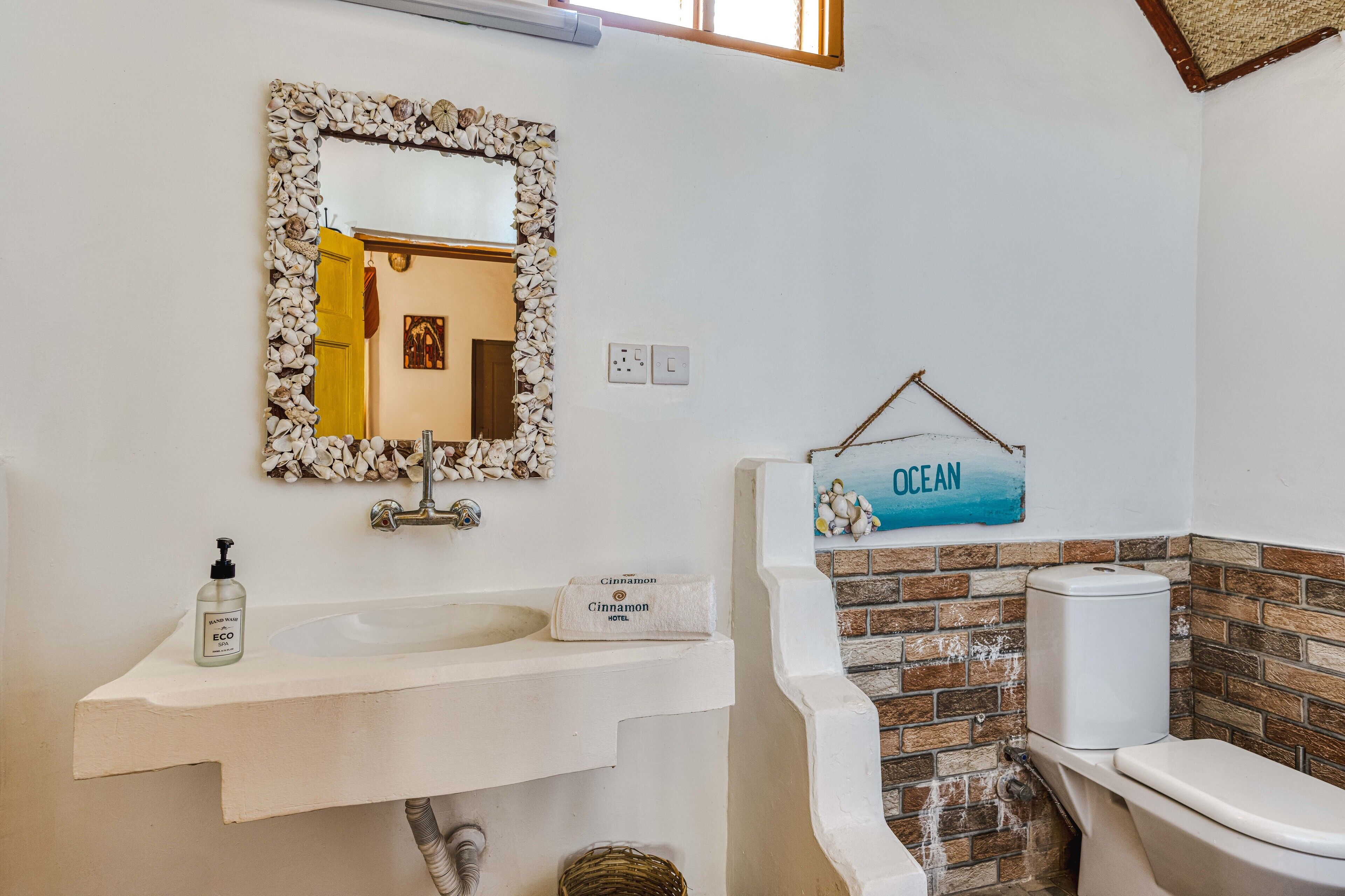 Une salle de bain avec un lavabo blanc, un miroir encadré de coquillages, des toilettes adossées à un mur en briques et un panneau indiquant « OCÉAN ».