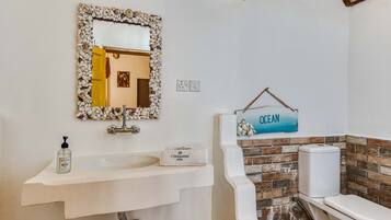 Une salle de bain avec un lavabo blanc, un miroir encadré de coquillages, des toilettes adossées à un mur en briques et un panneau indiquant « OCÉAN ».