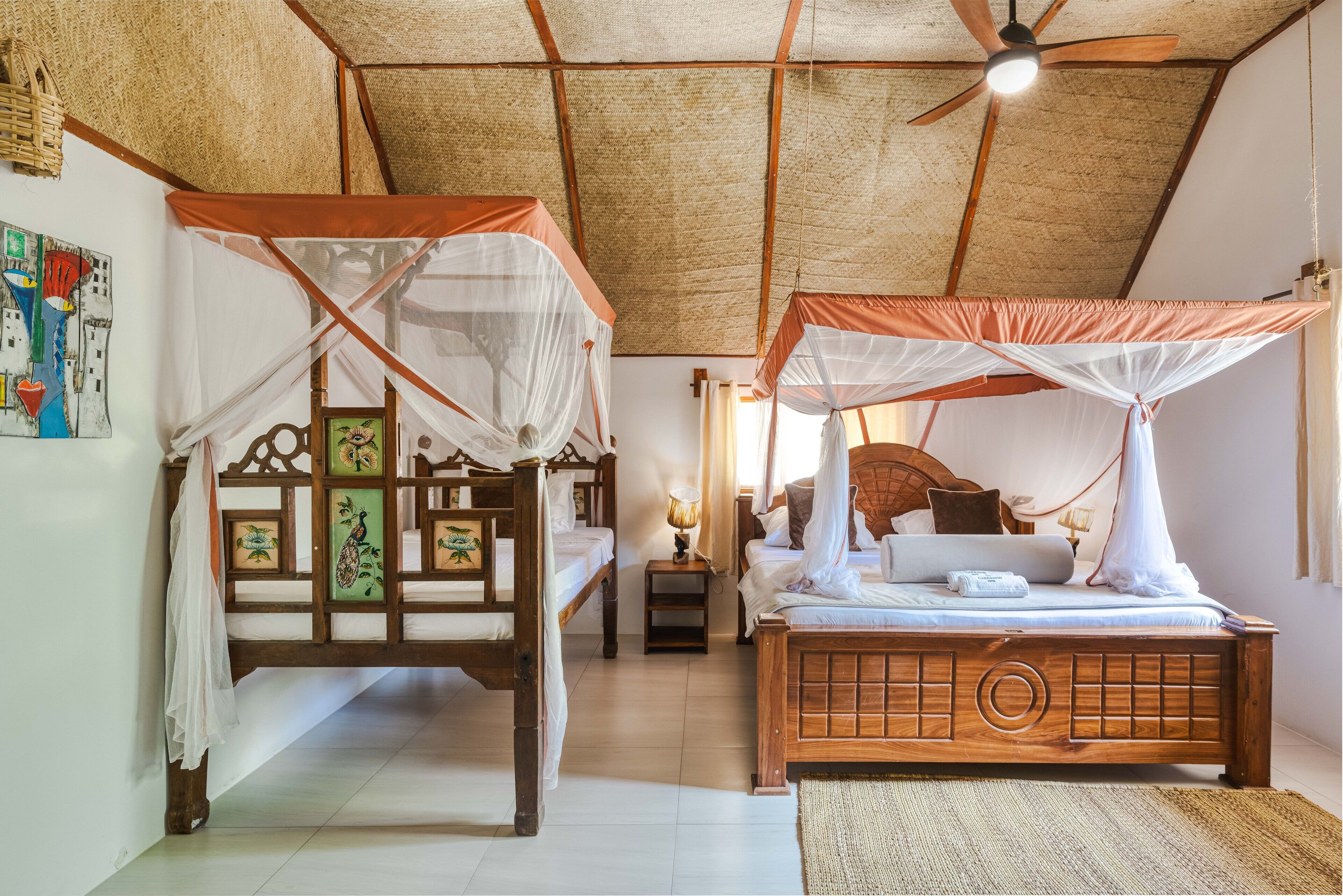 A traditional room with two wooden beds, each with a canopy. There is a ceiling fan, a small table with a lamp, and a woven mat on the floor.