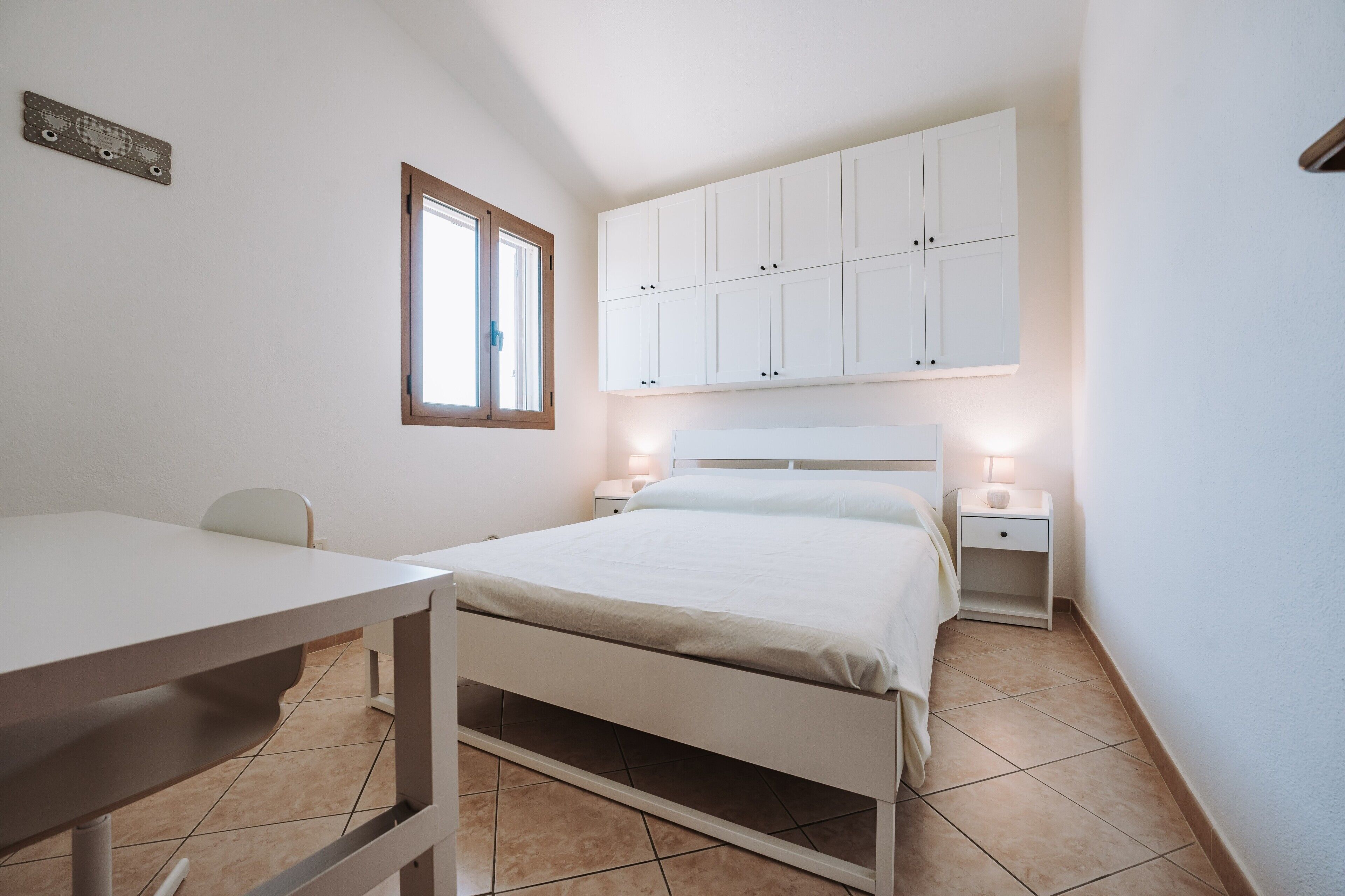 A bedroom with a bed, a desk, a chair, a wardrobe, and a nightstand with a lamp.