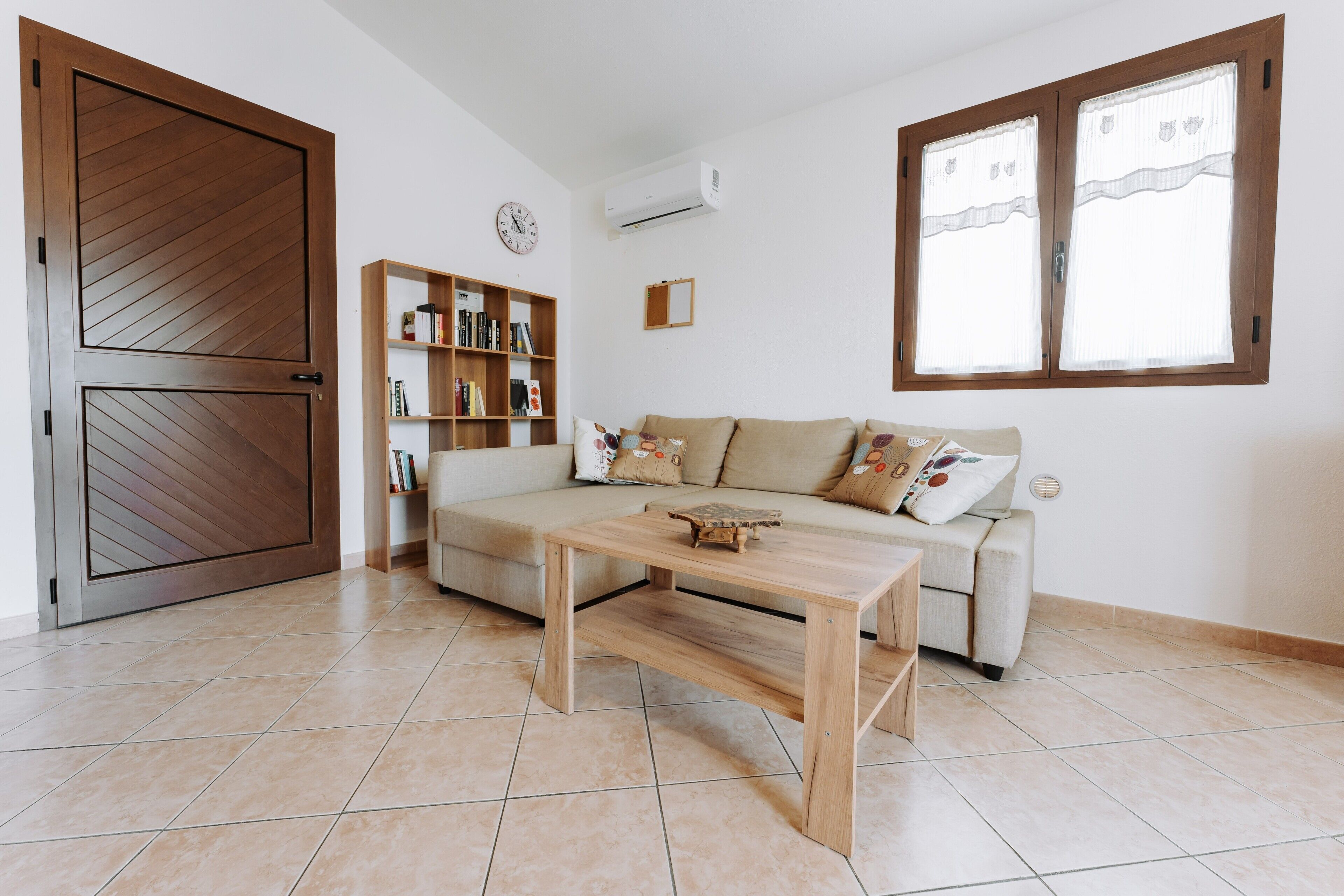 A living room with a sofa, a wooden coffee table, a bookshelf, and a large window with curtains.