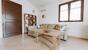 Una sala de estar con un sofá, una mesa de centro de madera, una estantería y un amplio ventanal con cortinas.