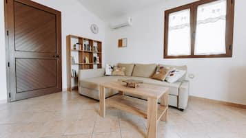 A living room with a sofa, a wooden coffee table, a bookshelf, and a large window with curtains.
