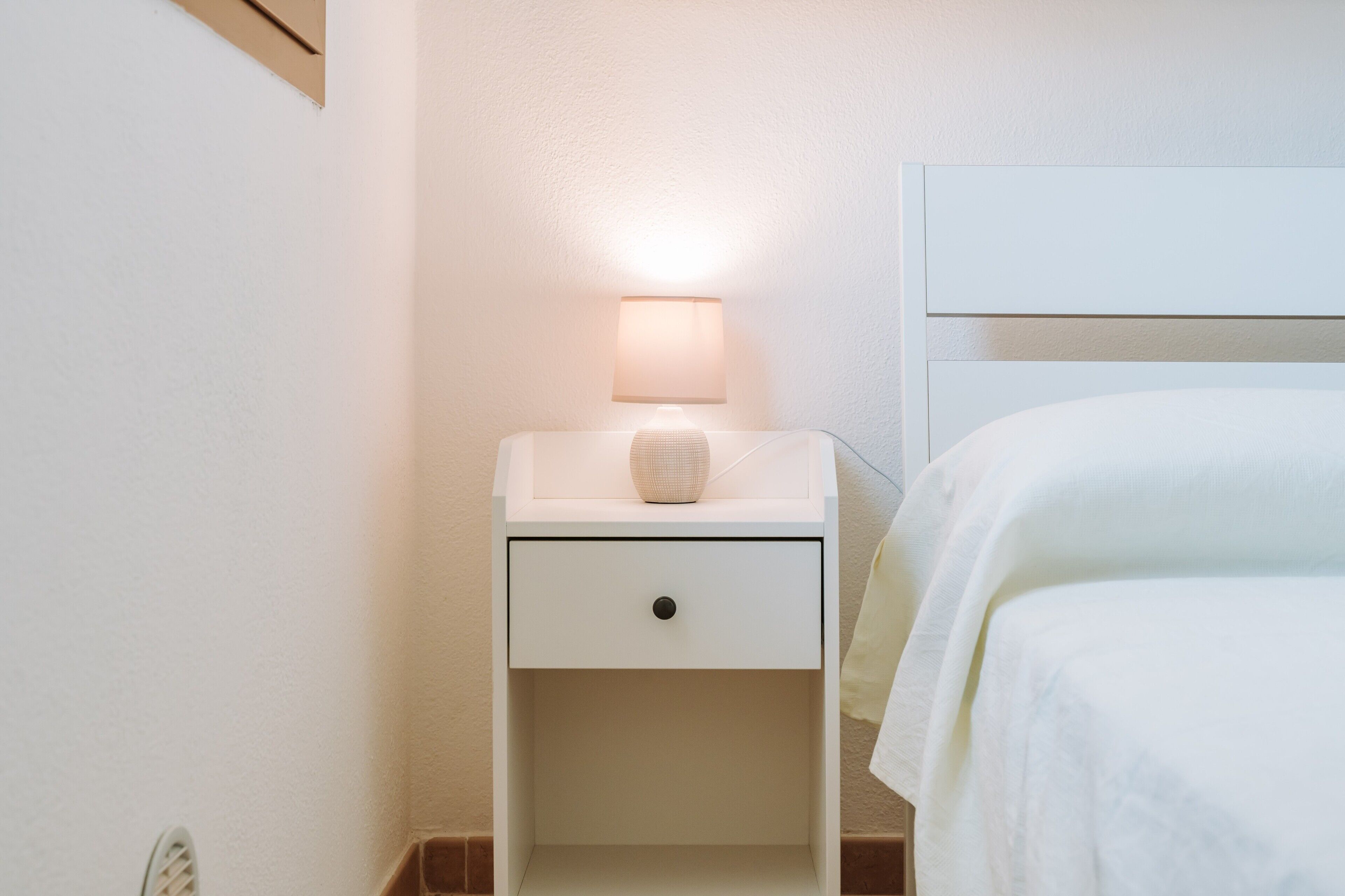 Une table de chevet blanche avec une lampe, un tiroir et un lit recouvert de linge de lit blanc.