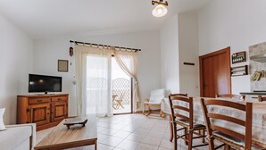 Una sala de estar con televisor, muebles de madera, una zona de comedor con sillas y una mesa, y una cocina con armarios y mostrador.