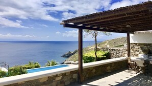 A stone patio with a pool, outdoor seating, and a view of the sea under a clear sky. - Mykonos Dot The Villas (Αλεόμανδρα)