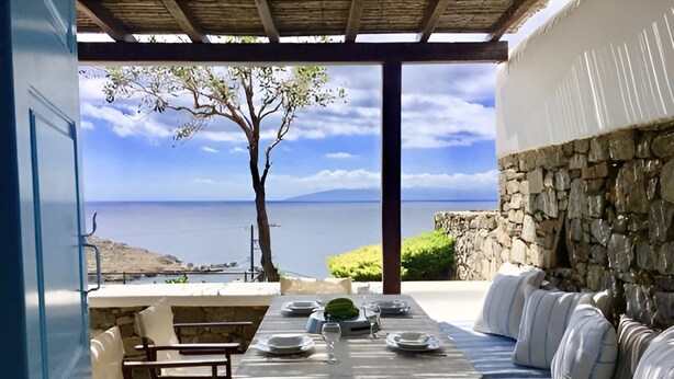 An outdoor dining area with a stone wall, a wooden table set with plates, and a view of the sea. - Mykonos Dot The Villas (Αλεόμανδρα)