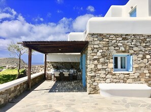 A stone building with a white exterior, a covered outdoor area with a table and chairs, and a view of the sea.