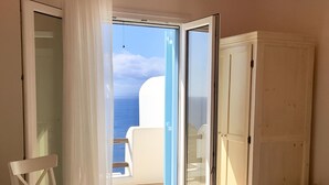 A bedroom with a bed, a chair, a wardrobe, and a sliding glass door leading to a balcony with a view of the sea. - Mykonos Dot The Villas (Αλεόμανδρα)