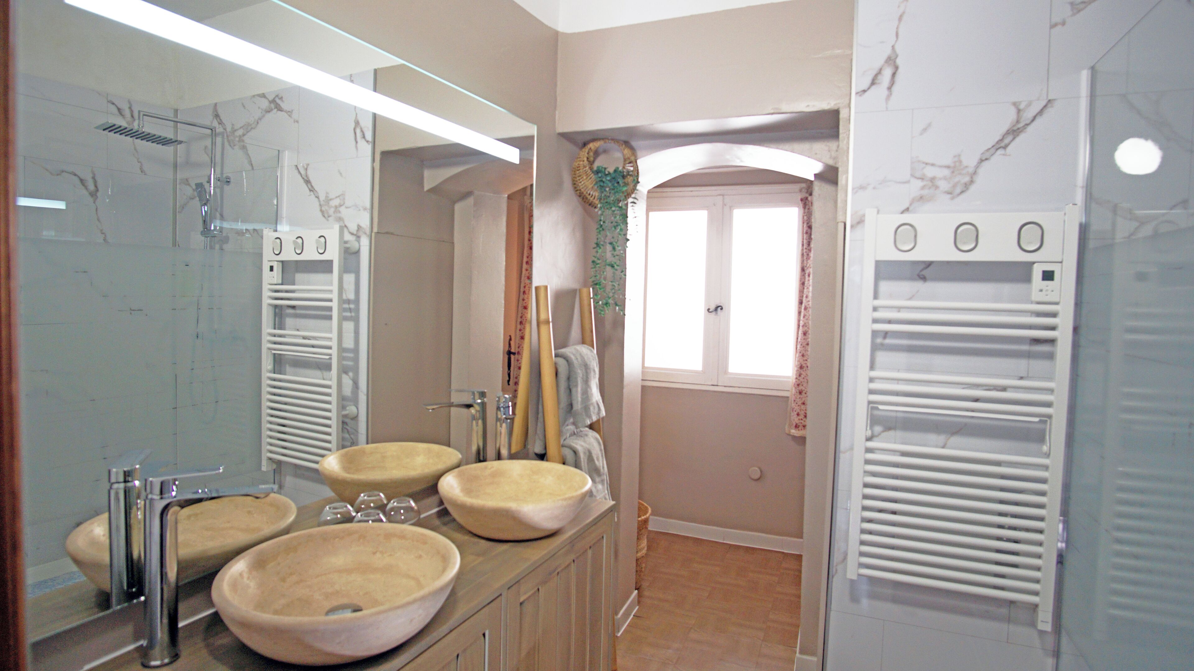 A bathroom with marble walls, a large mirror, two sinks, a shower, and a towel rack.
