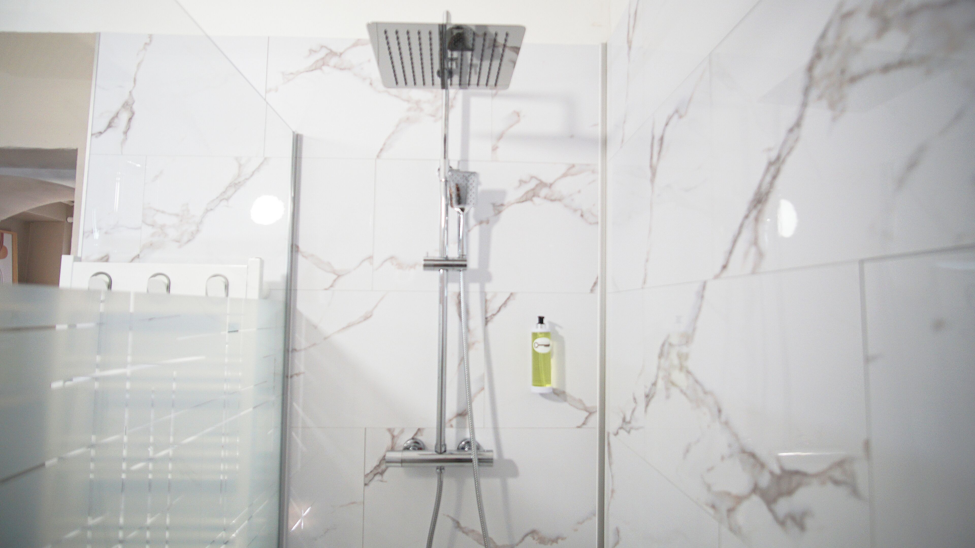 Een badkamer met douche, marmeren wanden en een fles shampoo.