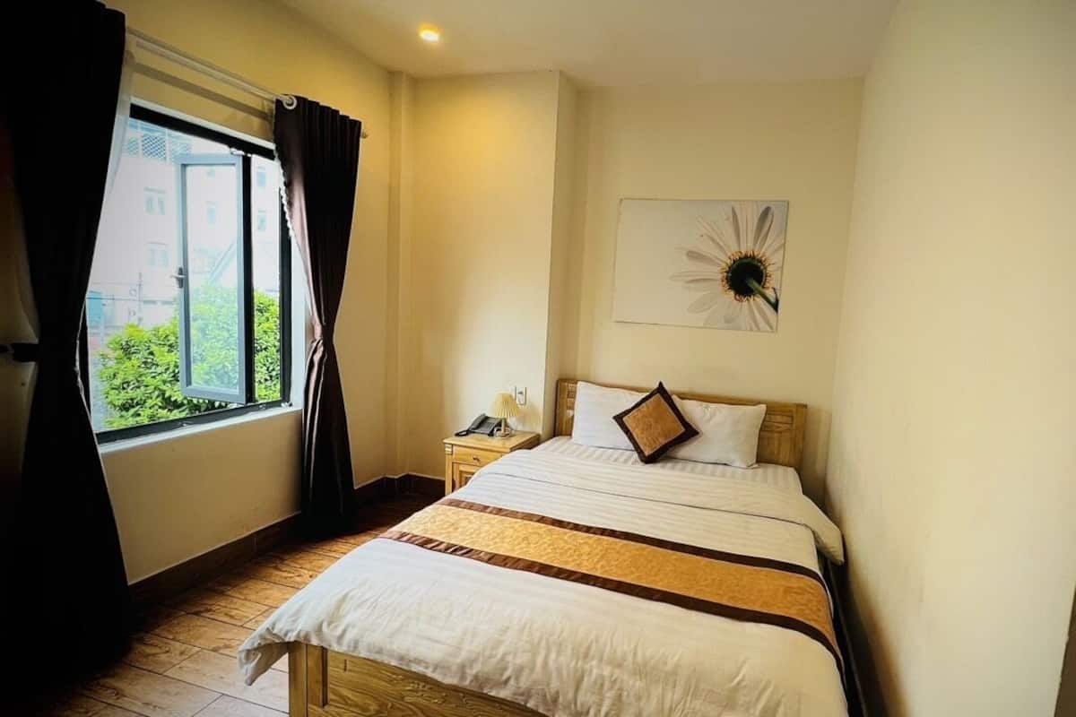 A hotel room with a bed, a nightstand, a window with curtains, a wall clock, and a picture of a sunflower.