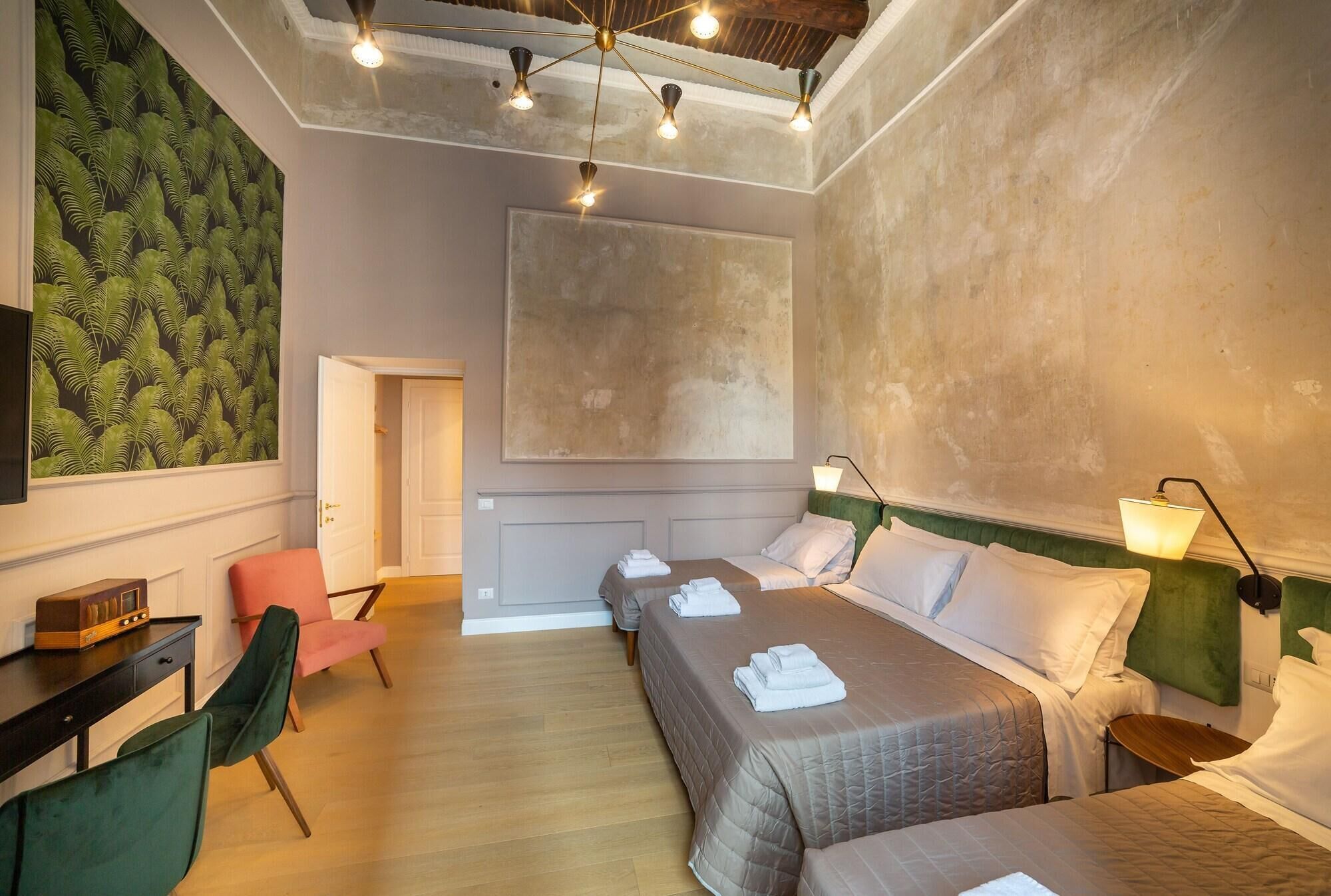 A hotel room with a large bed, two smaller beds, a desk with a chair, a television, and a green wall with a leaf pattern.