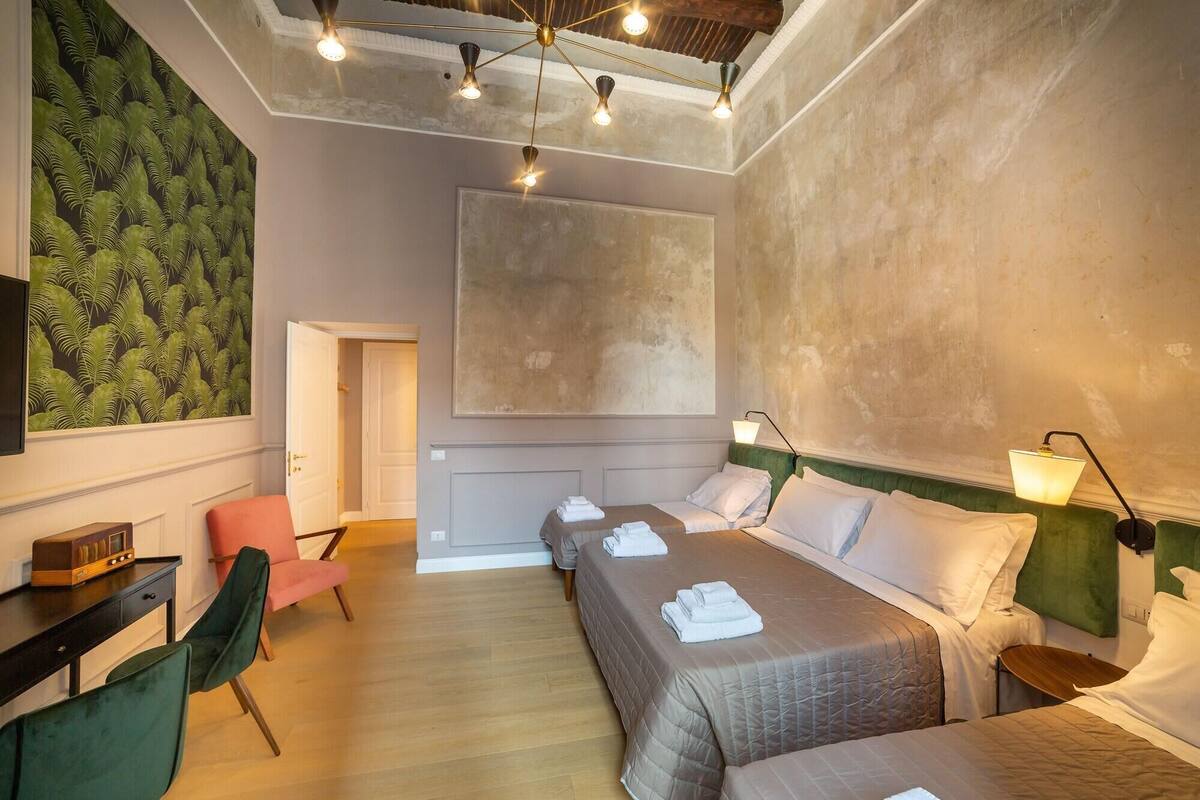 Une chambre d’hôtel avec un grand lit, deux petits lits, un bureau avec une chaise, une télévision et un mur vert orné d’un motif de feuilles.